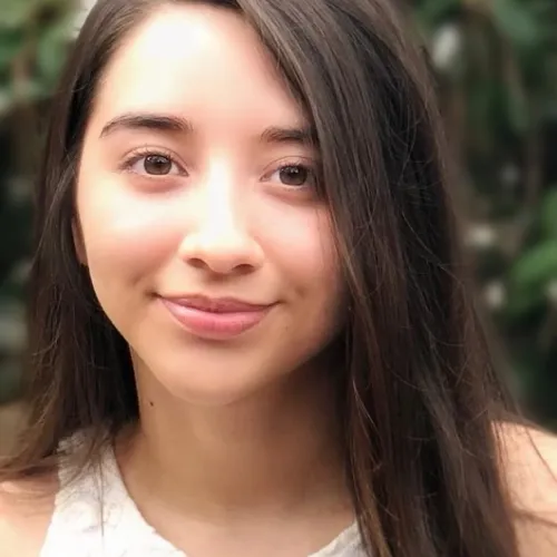 A close-up of a young woman with medium skin tone, straight dark brown hair parted to the side, wearing a white top with a lace pattern. She is smiling slightly and looking directly at the camera. The background is softly blurred with green foliage.