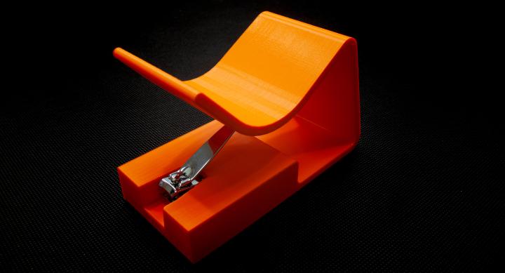 Orange nail clipper holder with slot to insert clippers at the bottom, and sloped force applicator up top.